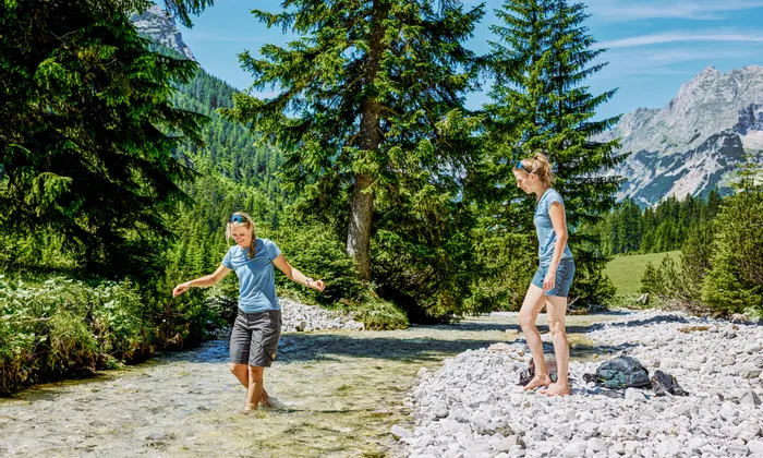 Zwei Frauen waten barfuß durch einen klaren Gebirgsbach inmitten von Wald und Alpenlandschaft